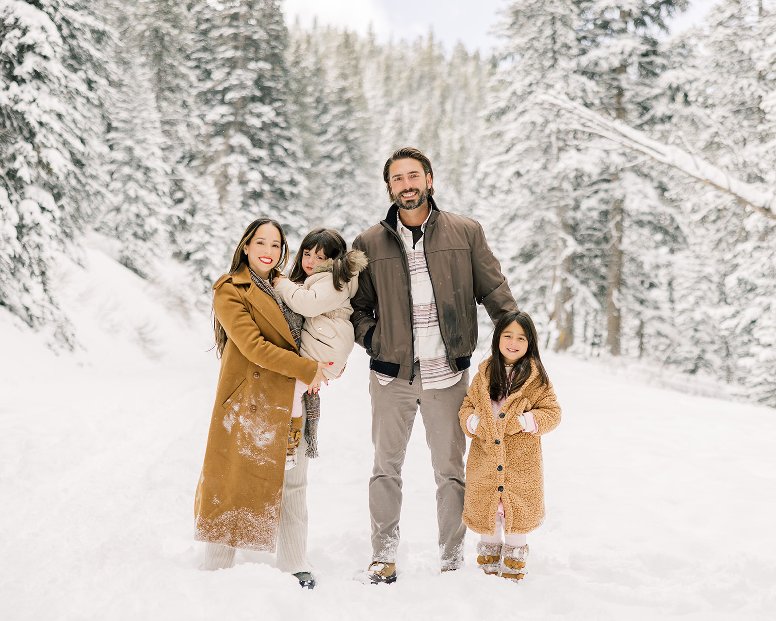 Happy mom and dad stand in a snow covered trail in a forest with their two young daughters in tan and brown coats