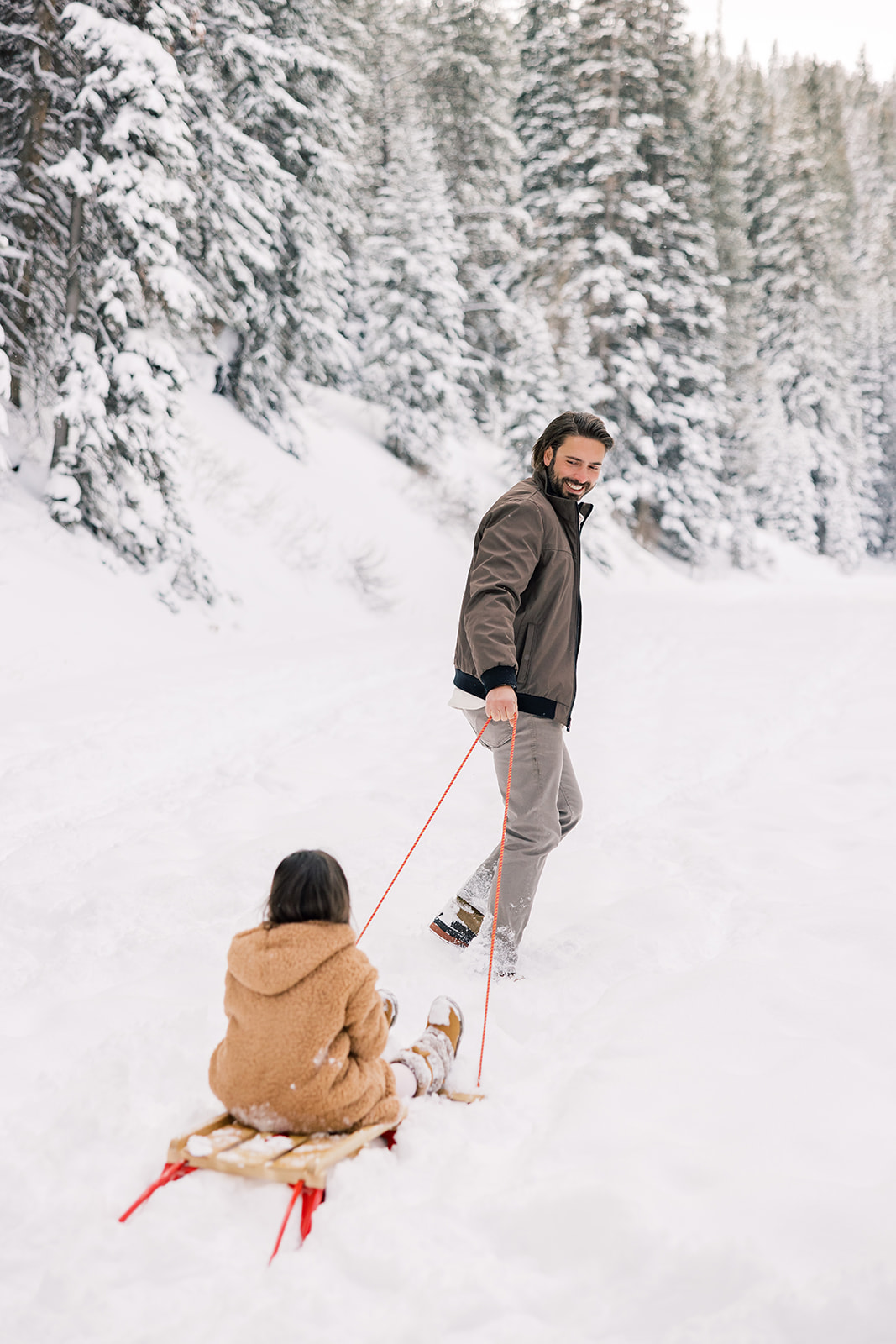 A happy dad pulls his toddler daughter on a sled through the snow in a forest after using Breckenridge Ski Resort Child Care