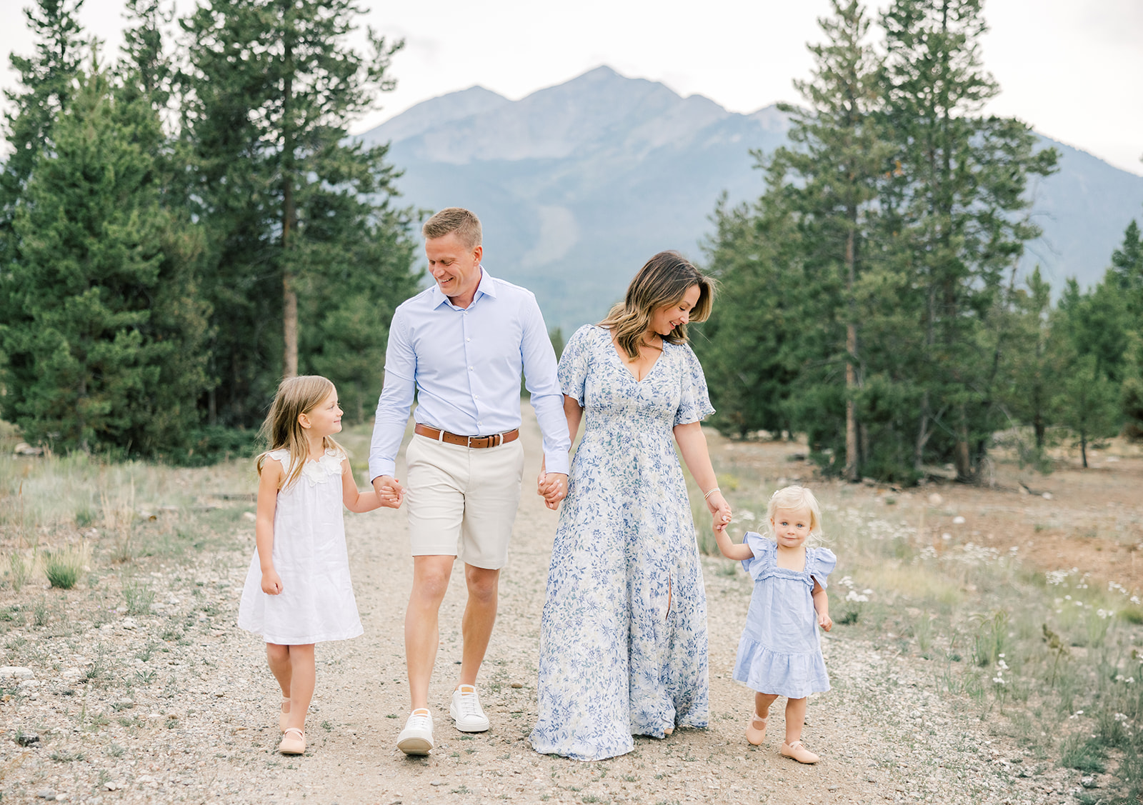 A mom and dad walk holding hands with their two toddler daughters in a mountain trail in blue outfits