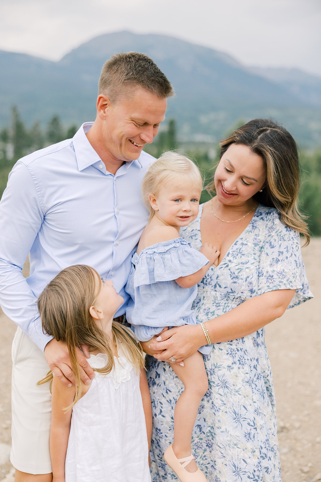 A happy mom and dad smile with their two toddler daughters while wearing blue in a mountain park after enjoying car services in Breckenridge