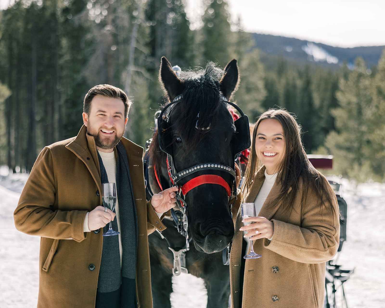Newly engaged couple hold champagne flutes while standing on a snowy mountain with a black horse