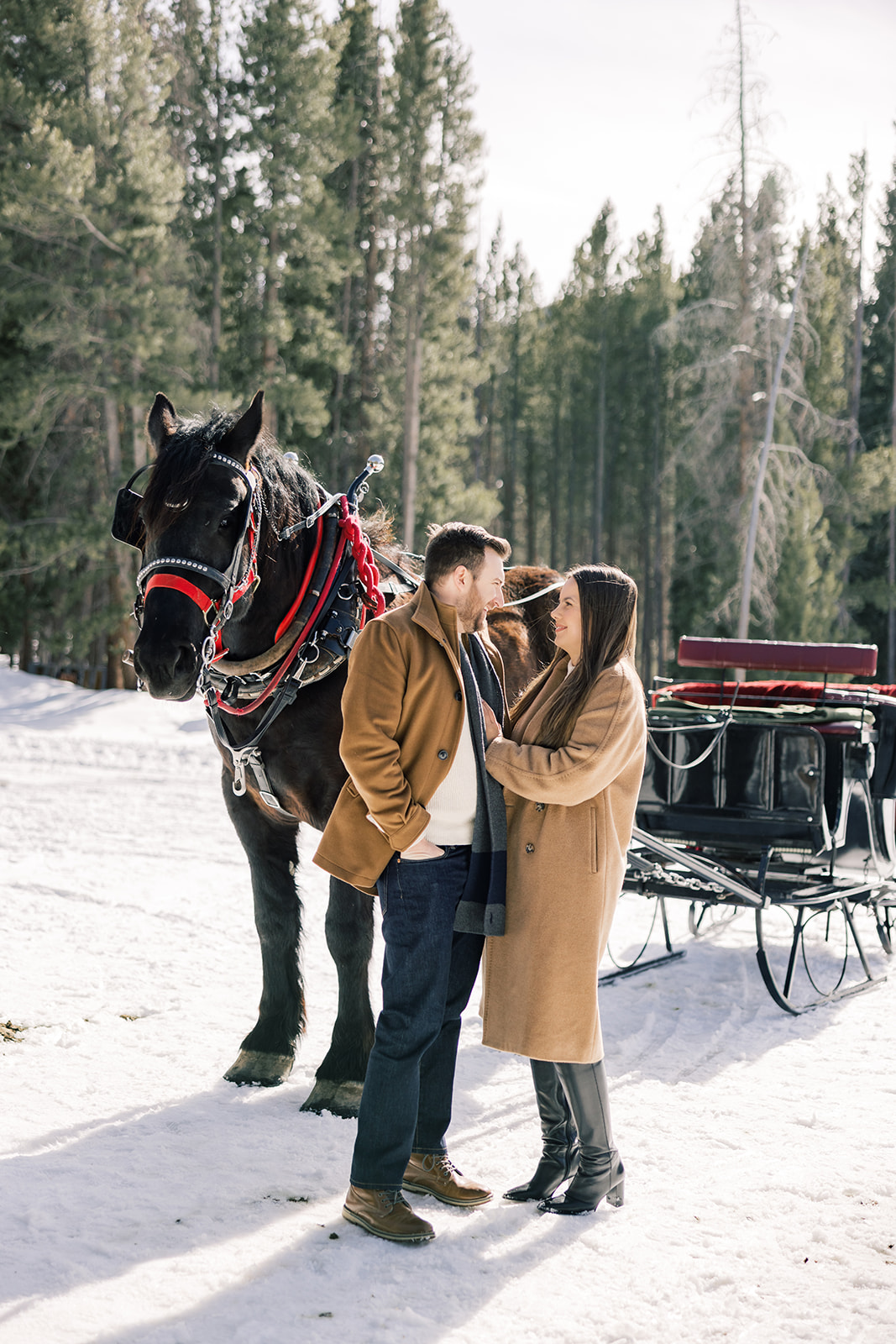 A happy couple in tan coats on a snowy mountain snuggle and smile at each other while on an adventure horseback riding in Breckenridge, CO
