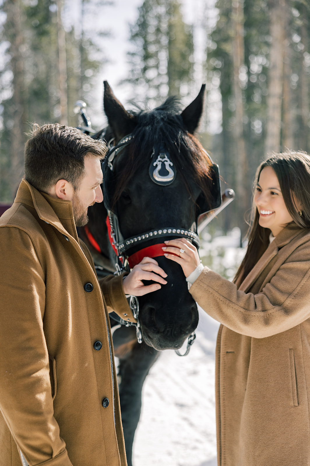 A happy couple in tan coats pet the nose of a horse while on an adventure horseback riding in Breckenridge, CO in the snow