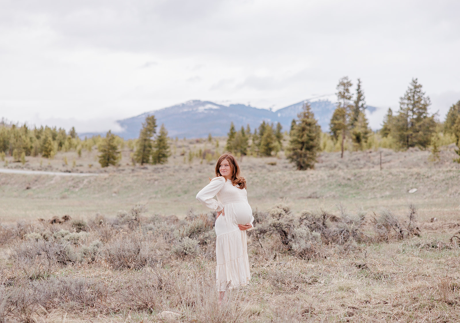 A pregnant woman rests her hand on her hip while standing in a meadow in a white maternity dress