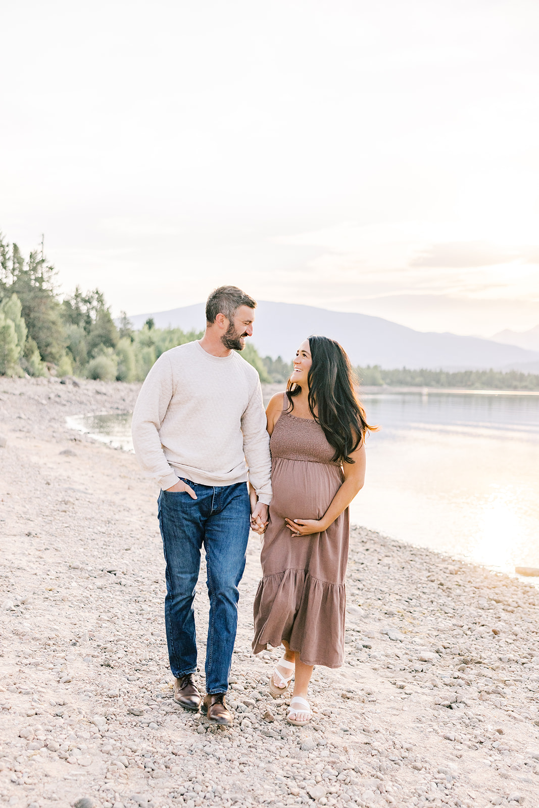 A mother to be walks along an alpine lake holding hands with her husband at sunset in a brown dress