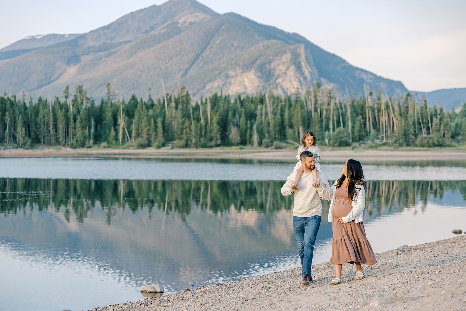 A happy mother to be walks along an alpine lake shore with her husband and toddler daughter on his shoulders in a brown maternity gown from Mama Bump Rentals