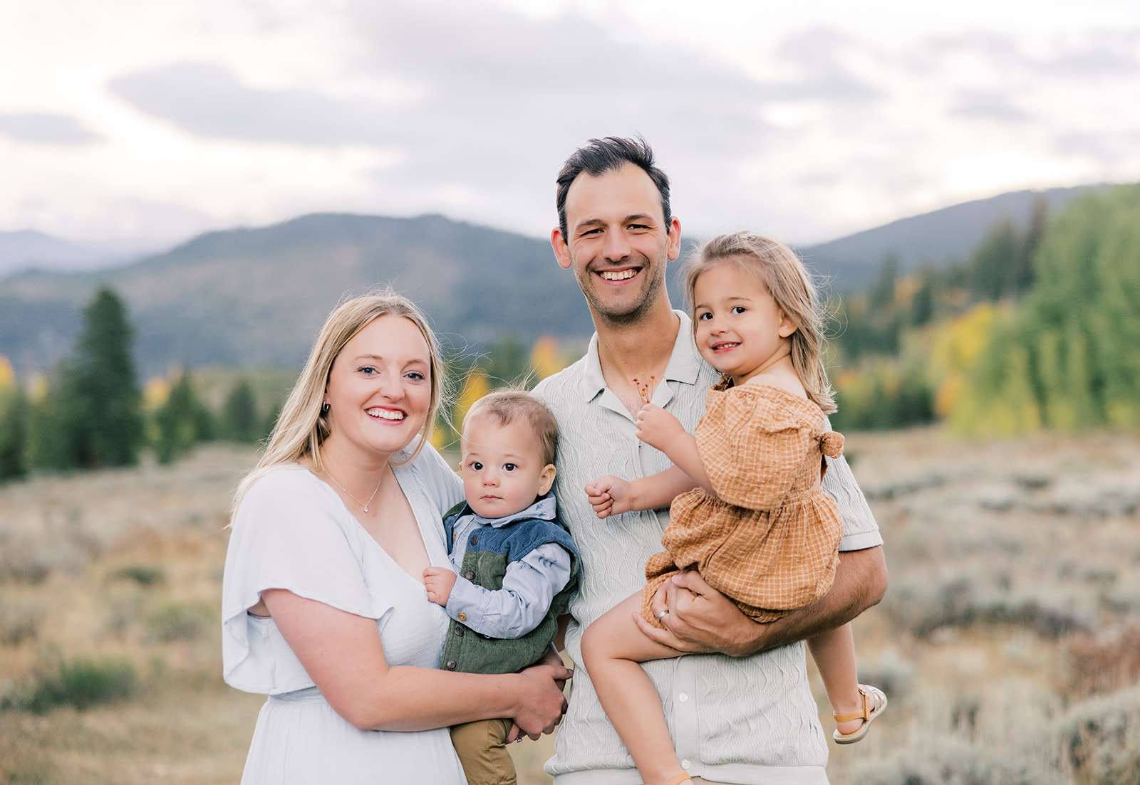 Happy mom and dad stand holding their toddler son and daughter on a hillside during guided hikes in Breckenridge