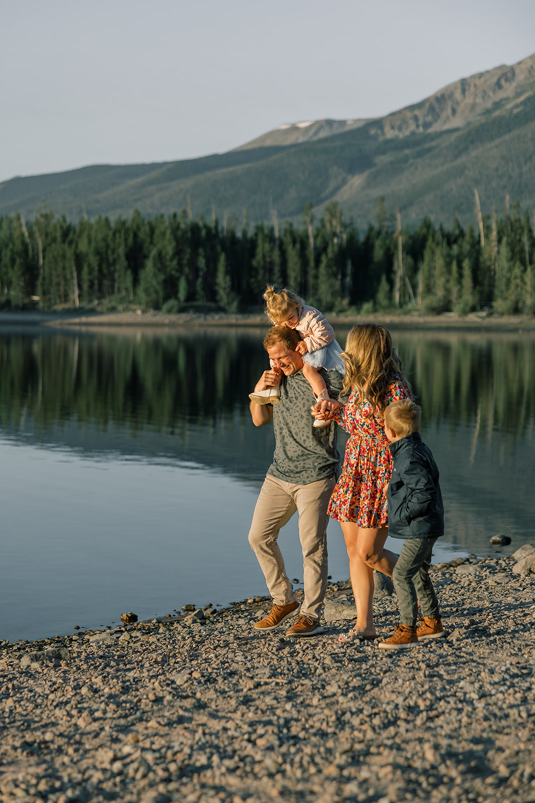 A happy mom and dad walk along an alpine lake shoreline holding their toddler daughter on dad's shoulders and hands with young son during some off-road tours in Breckenridge, CO