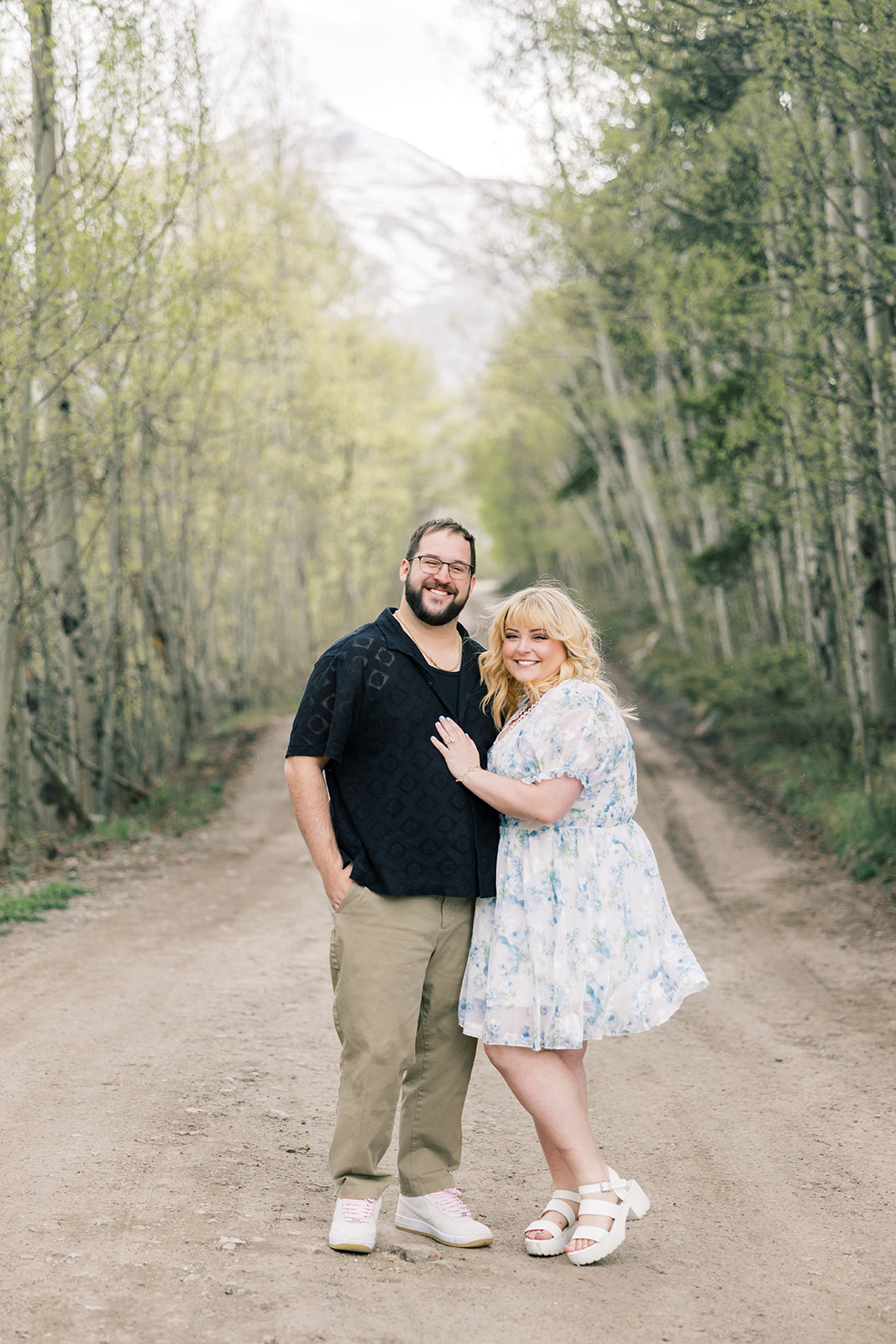 A happy couple stand in a forest dirt road leaning on each other and smiling