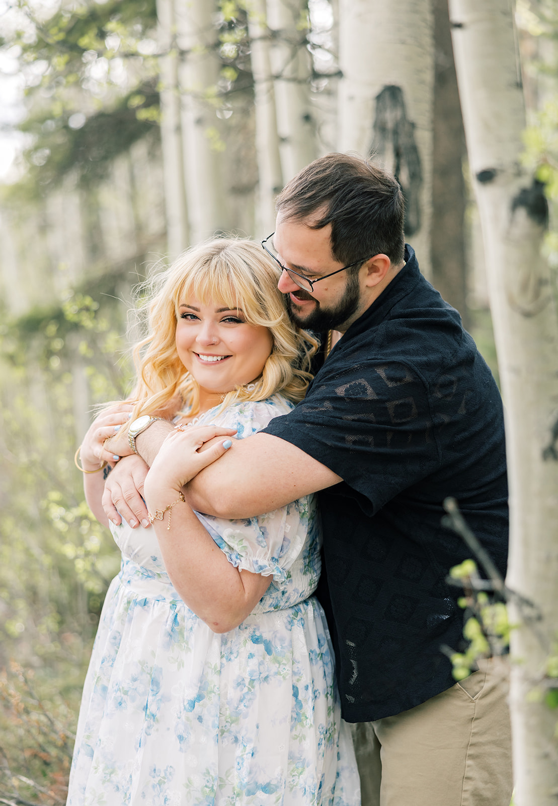 A happy woman in a blue floral print dress smiles while leaning into the chest of her partner in a black shirt in an. aspen forest while staying at luxury cabins in Breckenridge, CO