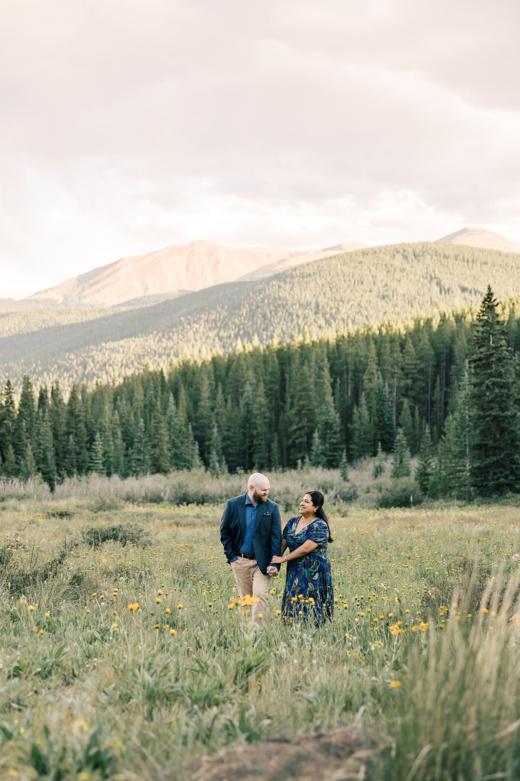 A happy couple walk holding hands through a meadow of yellow wildflowers in blue outfits at sunset after enjoying private dining in Breckenridge