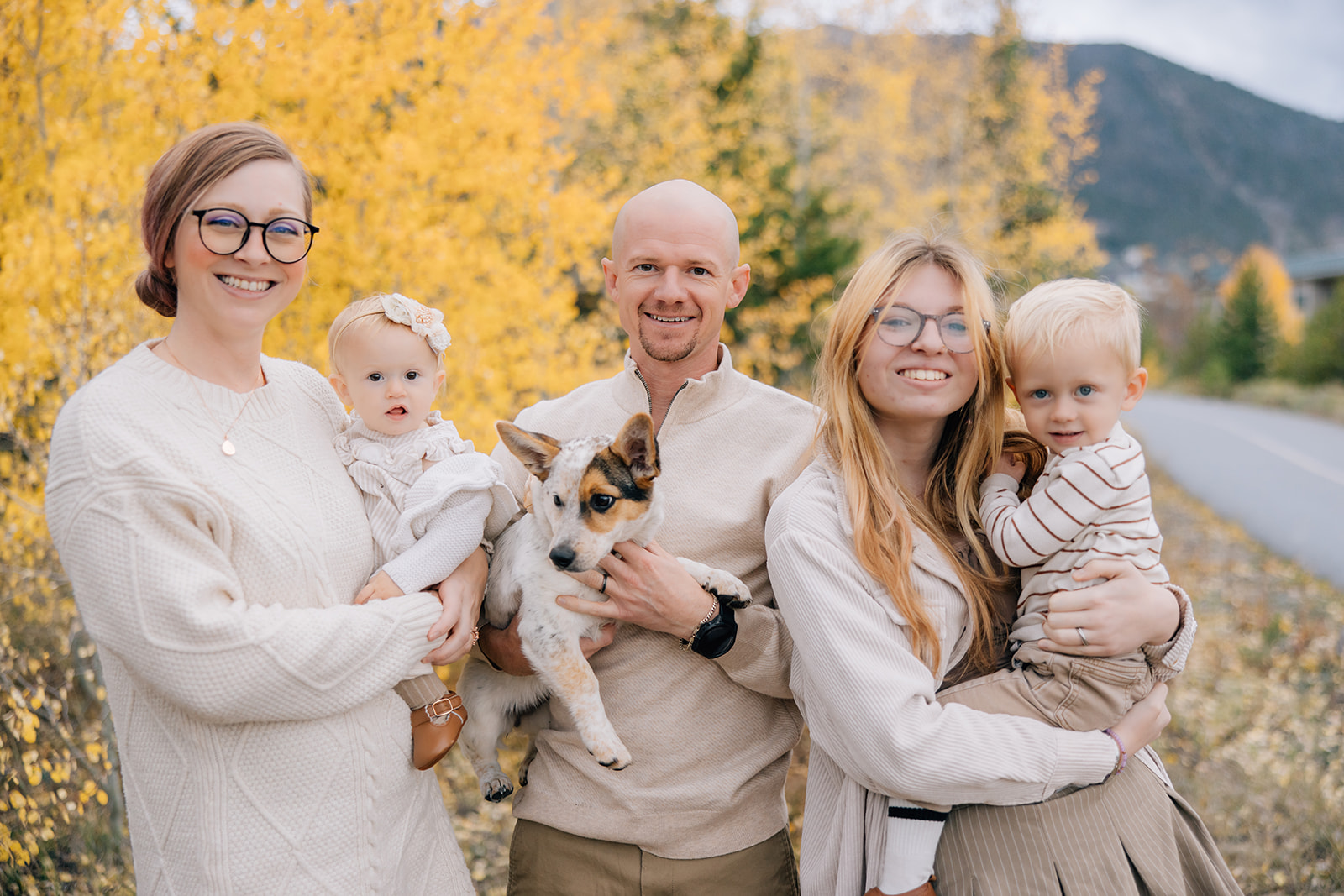 Three adults smile while standing in an autumn forest holding two toddlers and puppy