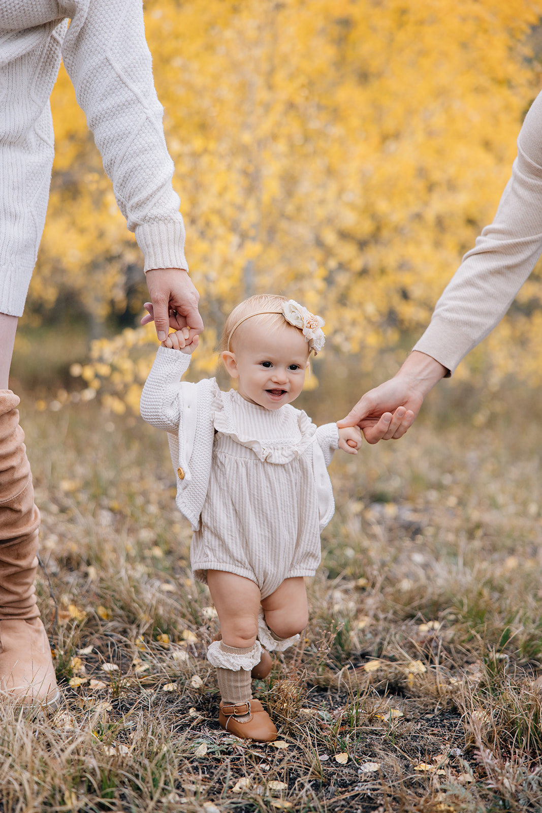 A toddler girl in a white onesie and sweater walks in a trail with help from mom and dad during one of the fall activities in Breckenridge, Colorado