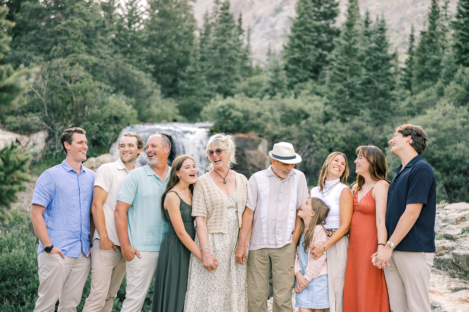 A happy large extended family laughs and holds hands while standing together near a waterfall before some whitewater rafting near Breckenridge, CO