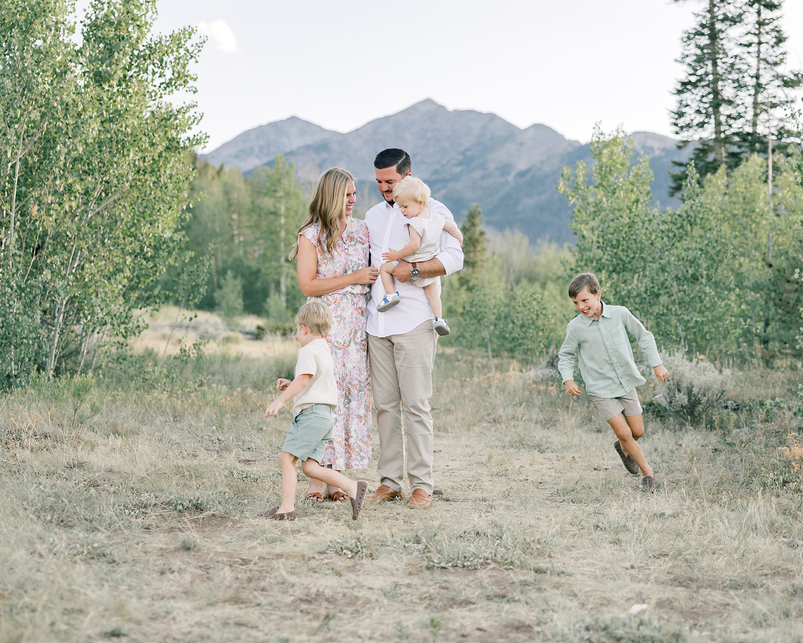 A mom and dad stand in a mountain trail as their two yhoung sons chase each other around them