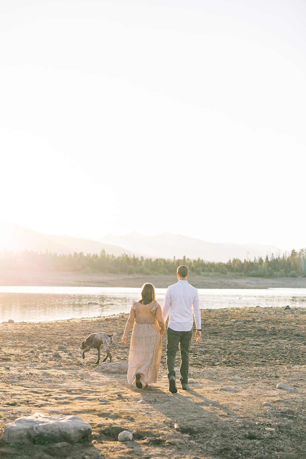 A happy pregnant couple walks holding hands with their dog on a riverbank at sunset during one of the unique things to do in Breckenridge