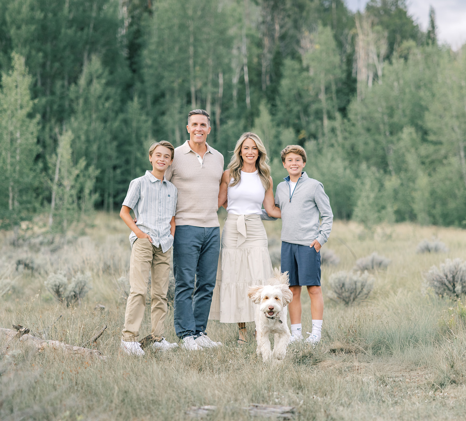 A happy mom and dad stand in a meadow with their two young sons and white doodle during one of the Breckenridge excursions