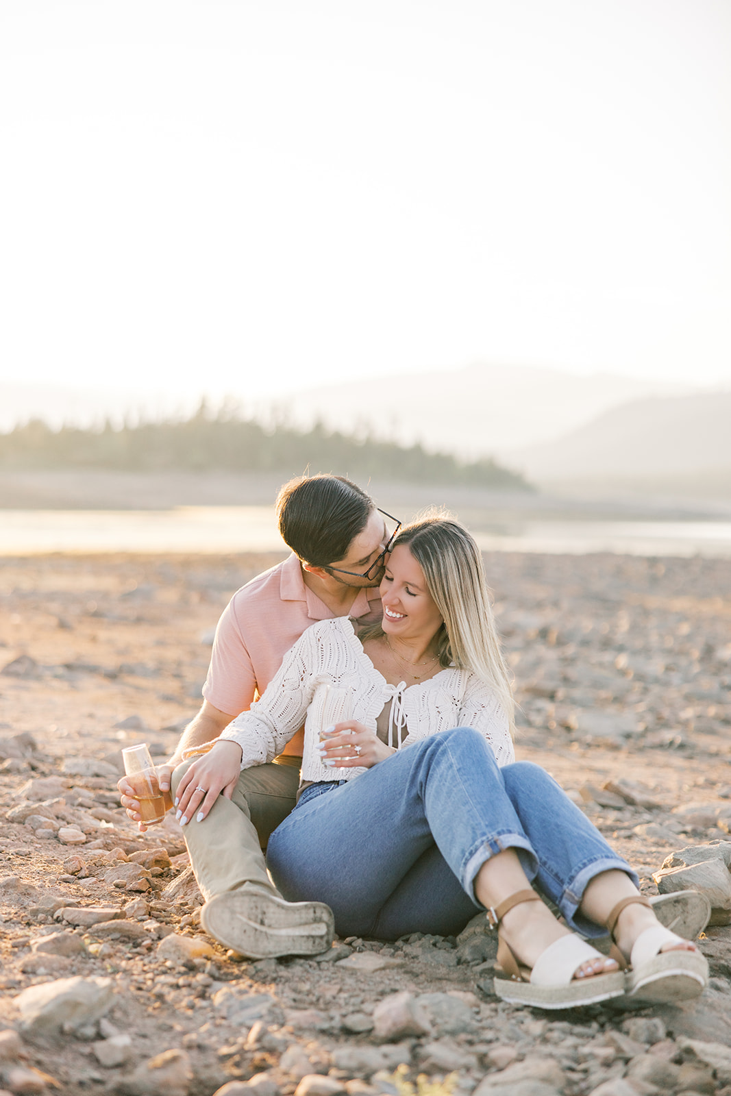 A happy newly engaged couple snuggles on the rocks near an alpine lake at sunset in white and pink
