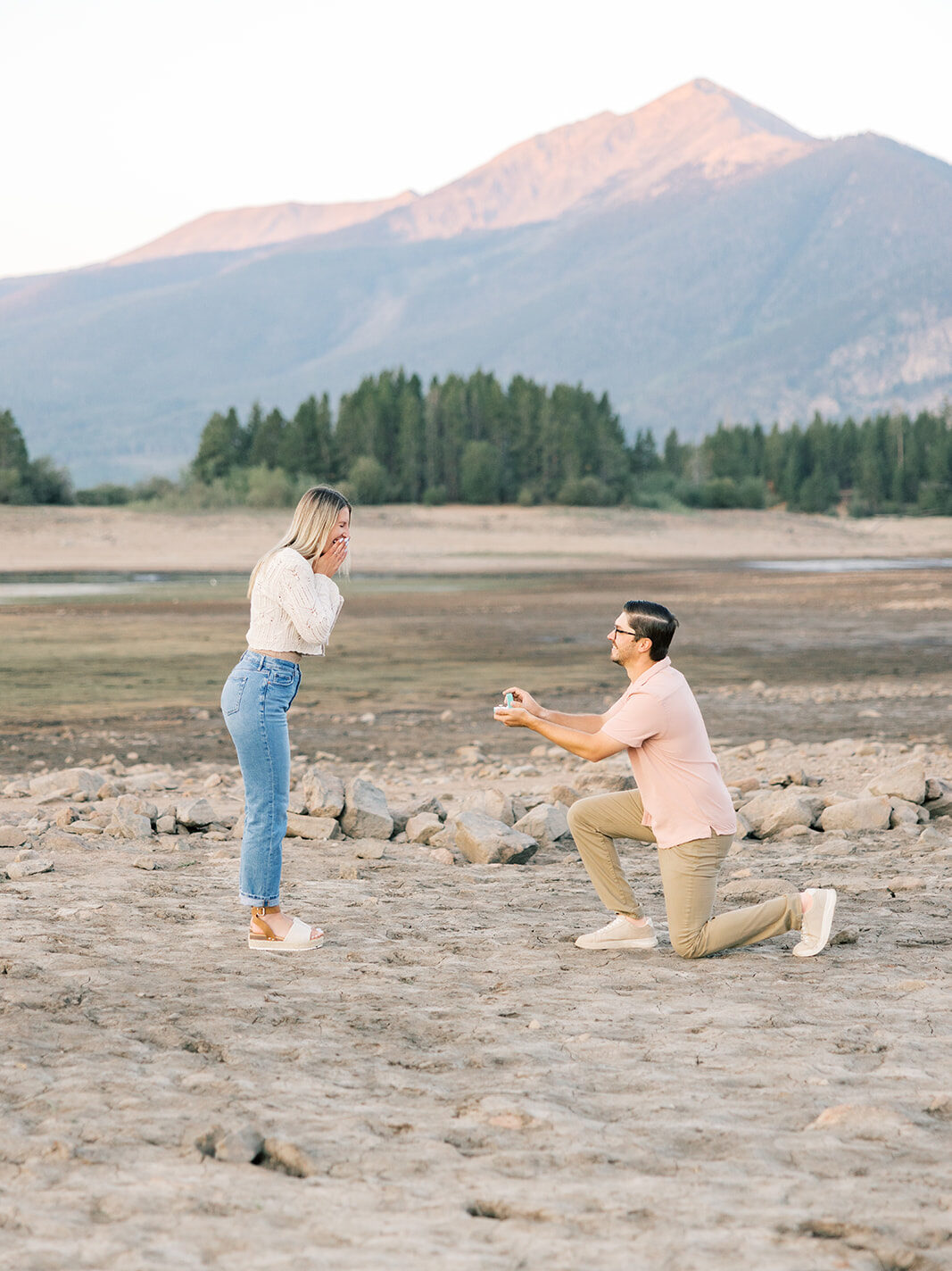 A man in a pink shirt gets on one knee to hold a ring box out for his girlfriend at one of the places to propose in Breckenridge at sunset