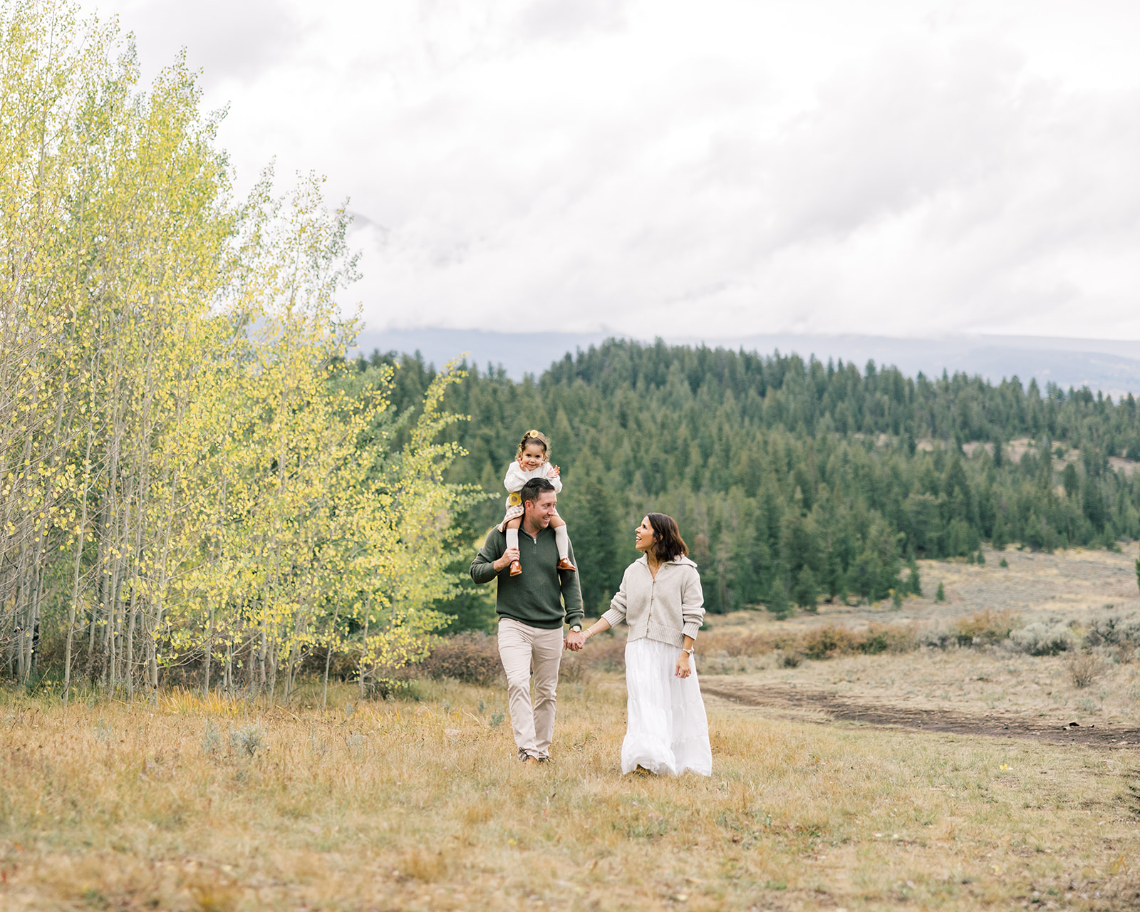 A mom walks holding hands with dad on a hillside with toddler on dad's shoulders while laughing
