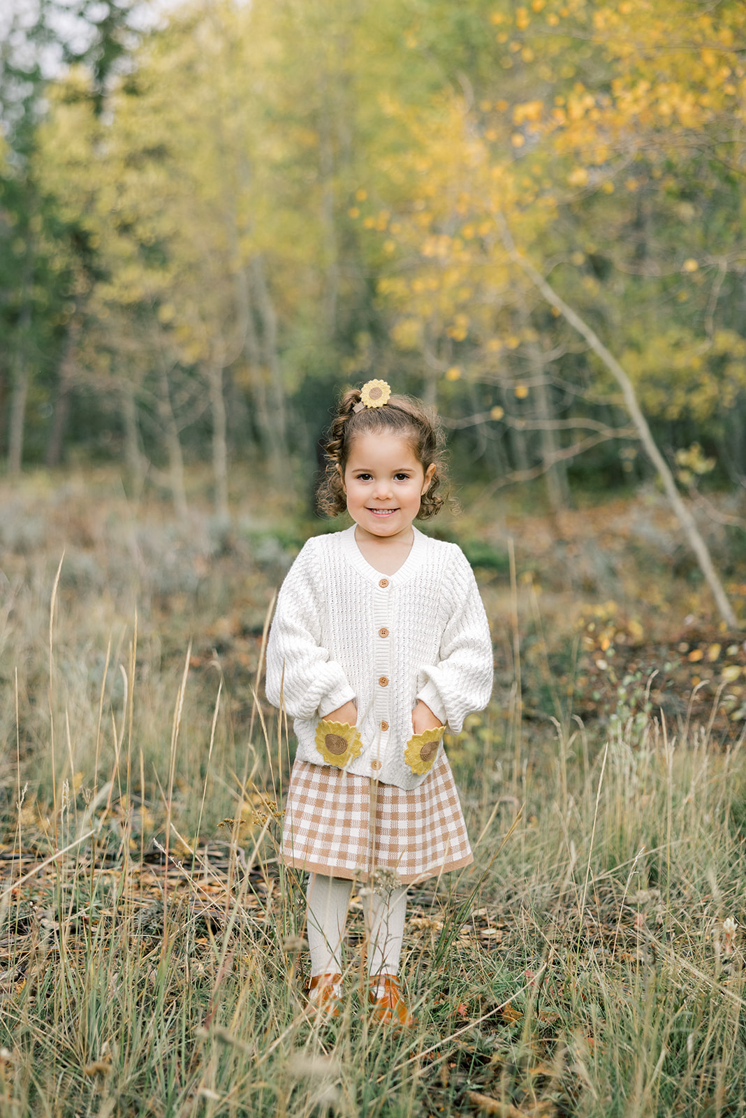 A toddler girl smiles with hands in her white sweater pockets in a forest in fall before finding indoor activities in Breckenridge