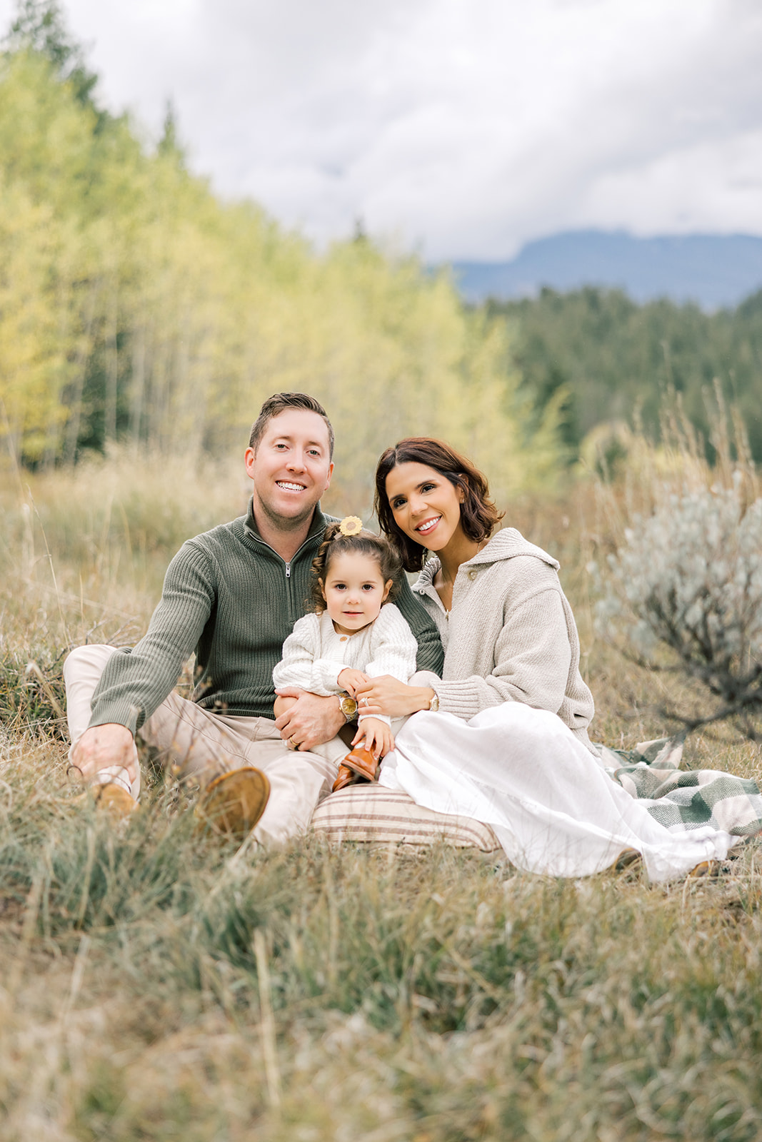 A toddler girl in a white sweater sits in mom and dad's laps in a mountain meadow before finding indoor activities in Breckenridge