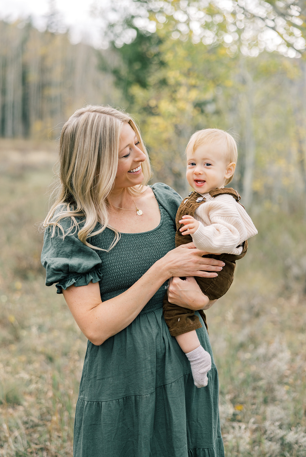 A happy mom in green dress stands in a forest trail smiling at her toddler son in her arms while enjoying long term rentals in Breckenridge, CO